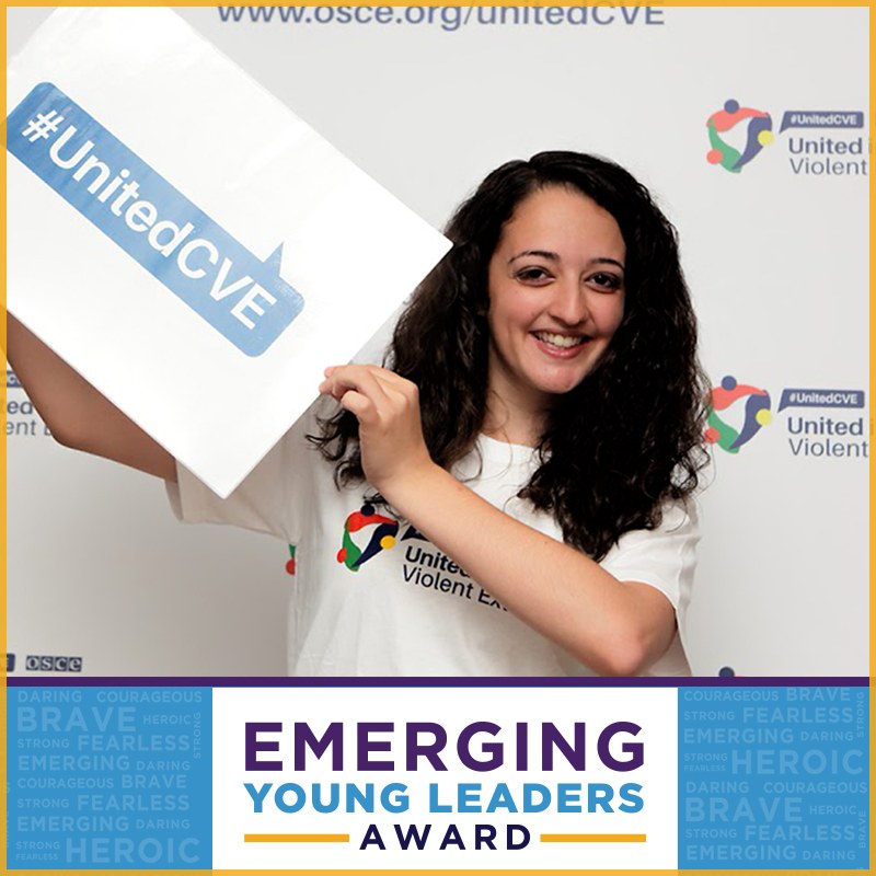Eine lachende Frau in einem weißen T-Shirt und Pferdeschwanz hält ein Schild mit der Aufschrift "Emerging Young Leaders Award" hoch und zeigt Freude und Errungenschaften.