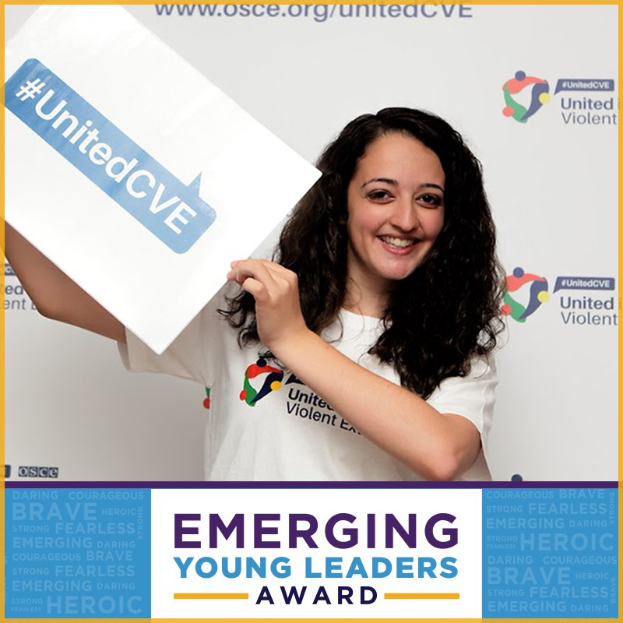 Eine lachende Frau in einem weißen T-Shirt und Pferdeschwanz hält ein Schild mit der Aufschrift "Emerging Young Leaders Award" hoch und zeigt Freude und Errungenschaften.