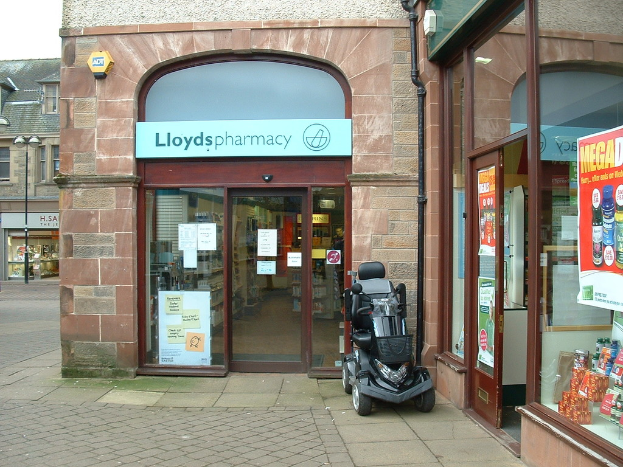 Eine Apothekenfiliale mit einem Fahrzeug davor und einem Gebäude links daneben.