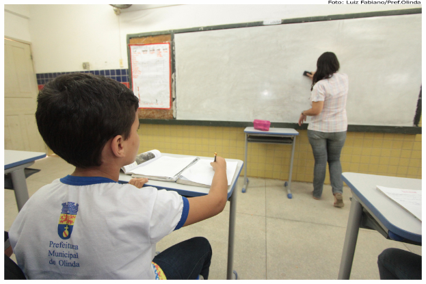 Ein Junge sitzt an einem Tisch und schreibt in ein Buch, während eine Frau die Tafel mit einem Putzlappen putzt in einem Klassenzimmer mit mehreren Tischen, einem Plakat und einer Kiste auf einem Tisch.