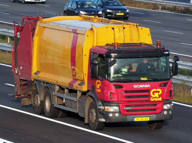 Ein gelb-roter Mülllaster fährt auf einer Autobahn, umgeben von anderen Fahrzeugen, mit einer Leitplanke und Gras auf beiden Seiten der Straße.