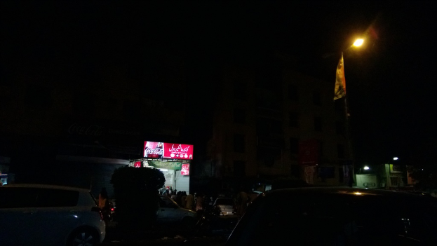 Eine nächtliche Szene einer Straße mit Autos, einem kleinen Laden im Hintergrund, einem Laternenmast auf der rechten Seite, einer Werbetafel über dem Laden und beleuchteten Gebäuden auf der rechten Seite.