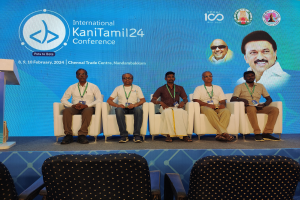 Eine Gruppe von Menschen, die vor einem Podium mit einem Mikrofon sitzen, mit einem Banner im Hintergrund mit Text und Bildern von Menschen und Stühlen unten, das die Internationale Kanitamil24-Konferenz suggeriert.