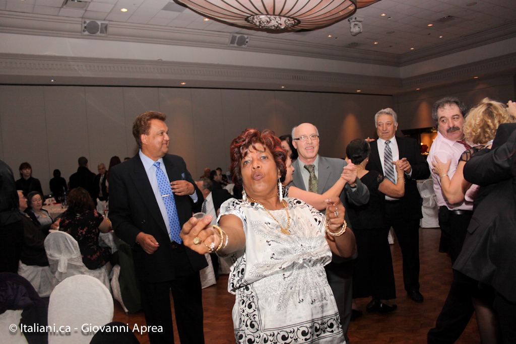 Eine Gruppe von Menschen, einige stehend und einige sitzend, in einem Raum mit einem Wasserzeichen auf dem Boden, Stühlen, einer Wand, Lampen und einem Lautsprecher, wobei eine Frau eine Halskette, Armbänder, Fingerringe und Ohrringe trägt.
