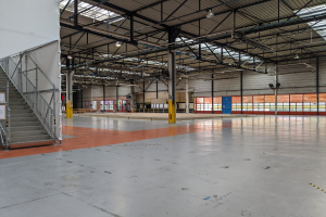 Großes Industriegebäude mit zahlreichen Fenstern, Metallträgern, einer Treppe mit Geländer, einem Schild, Deckenleuchten und zusätzlichen Pfählen.