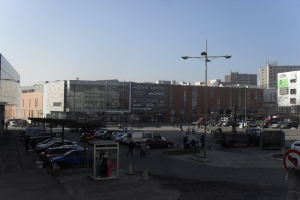 Eine belebte Stadtstraße mit einem Parkplatz voller Autos, hohen Gebäuden, Straßenlaternen und ein paar Passanten unter einem sichtbaren Himmel.