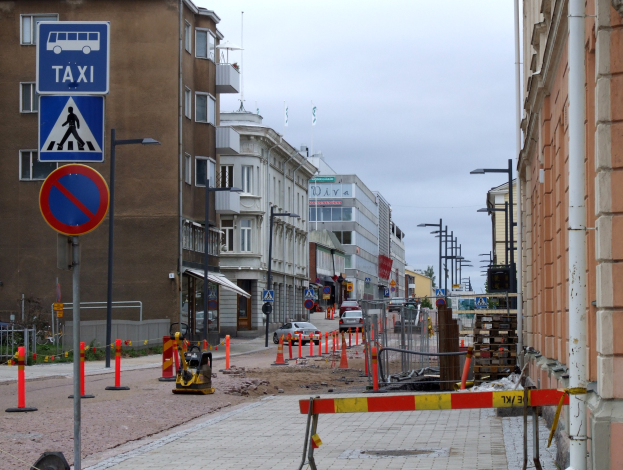 Eine Stadtstraße mit Gebäuden, Straßenlaternen, Verkehrsschildern, Verkehrszeichen, Baustellenschildern, Autos, Absperrpoller, Bäumen und einem Himmel mit Wolken im Hintergrund.