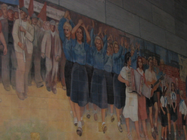 Eine Gruppe von Menschen mit blauen Mädchen in der Mitte, die klatschen, Musiker auf der rechten Seite, spielende Kinder in der rechten Ecke und Personen mit roten Fahnen auf der linken Seite.