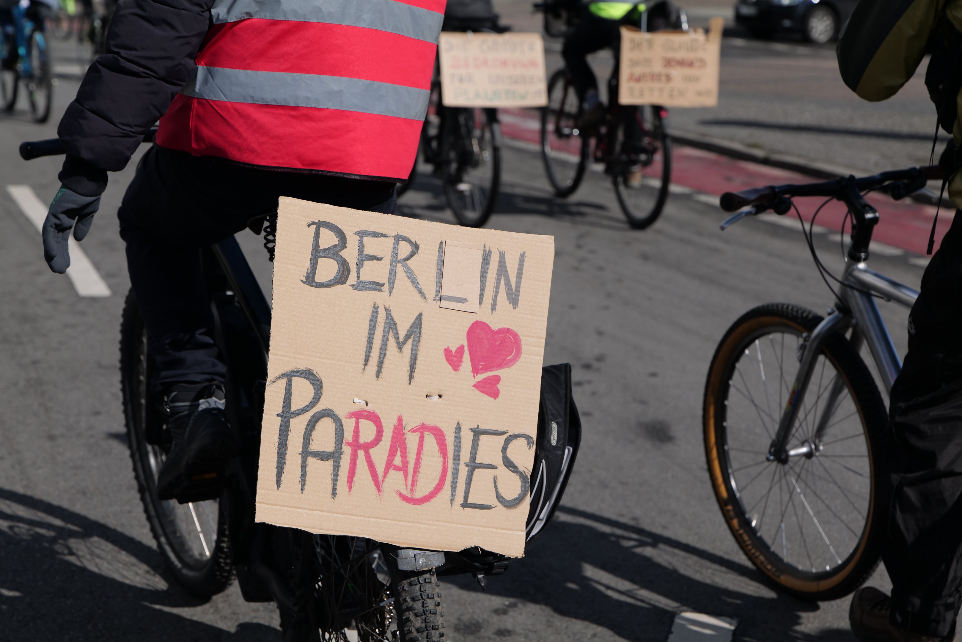 Eine Gruppe von Menschen, die auf Fahrrädern die Straße entlangfahren, mit einem "Berlin I'm Paradies"-Schild im Vordergrund und einem Auto im Hintergrund.