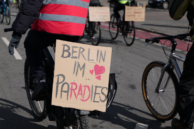 Eine Gruppe von Menschen, die auf Fahrrädern die Straße entlangfahren, mit einem "Berlin I'm Paradies"-Schild im Vordergrund und einem Auto im Hintergrund.