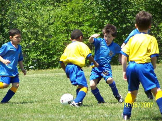 Kinder beim Fußballspielen auf einem grünen Feld mit Bäumen im Hintergrund und einem Datumsstempel in der rechten unteren Ecke.