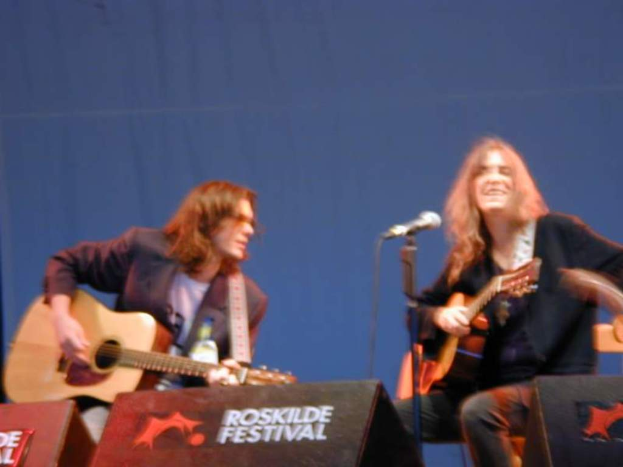 Zwei Männer auf der Bühne mit musikalischen Instrumenten, ein Mikrofon davor und zwei Kisten auf dem Boden mit 'ROCK SLIDE FESTIVAL' darauf.