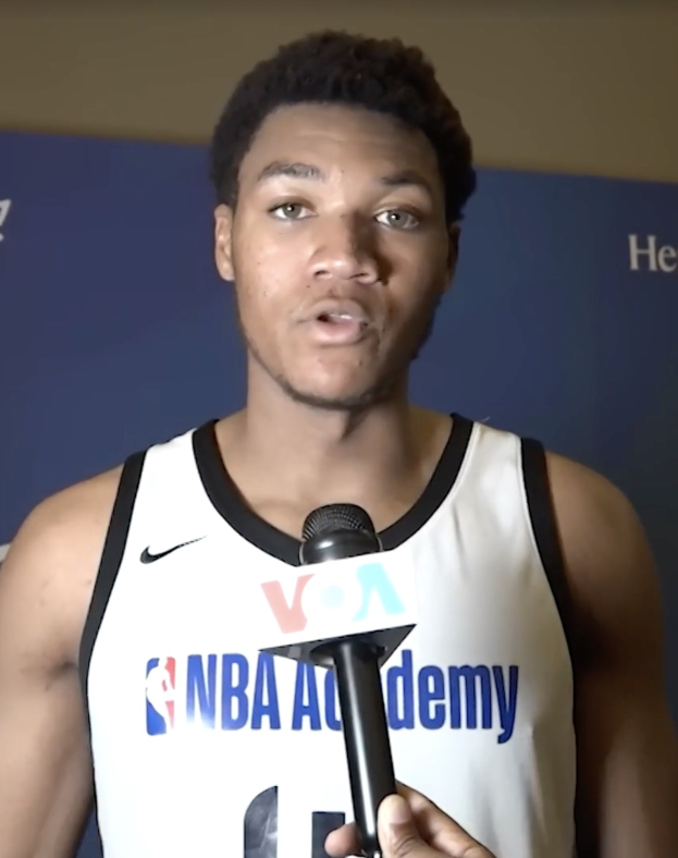 Mann in weißem Tanktop mit Mikrofon spricht mit der Presse bei der NBA All-Star-Game 2019, mit einem Banner und einer Wand im Hintergrund.