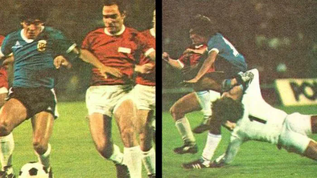 Eine Gruppe von Männern beim Fußballspielen auf einem Feld, wobei einer den Ball tritt und andere versuchen, die Kontrolle zu übernehmen, mit einer Tafel im Hintergrund, die auf eine Stadionatmosphäre hindeutet.