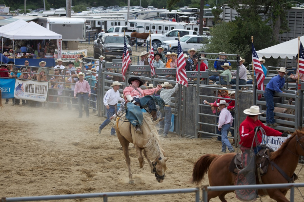 Zwei Reiter auf Pferden bei einem Wettbewerb namens Charreada, umgeben von Gruppen von Menschen, Banner und Fahnen, Pavillons, geparkten Fahrzeugen und Bäumen im Hintergrund.
