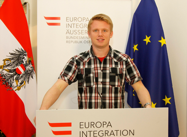 Ein Junge steht hinter einem Podium mit Mikrofon, mit einem Plakat und Fahnen dahinter.