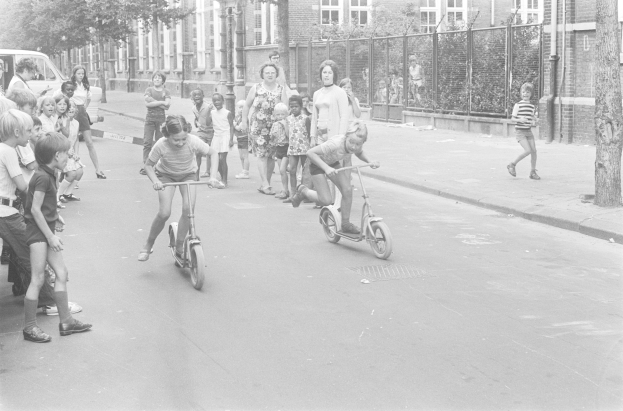 Schwarzes Bild von Kindern, die auf Roller die Straße entlangfahren, mit einem geparkten Auto auf der linken Seite, Gebäuden, Bäumen, Pfählen und einem Zaun im Hintergrund.