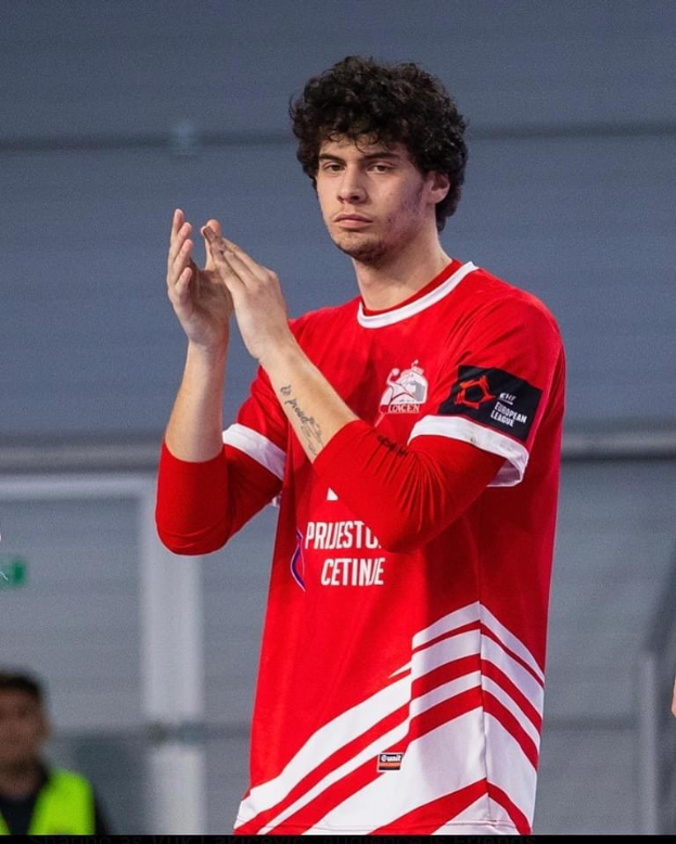 Ein Mann in einem roten und weißen Hemd klatscht in den Händen in den Vordergrund, mit einer anderen Person und einer Wand im Hintergrund und dem Text 'Kopenhagen Futsal Club - Foto 1' unten.
