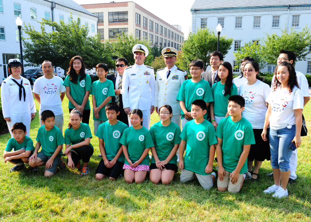 Eine Gruppe von Menschen in grünen Hemden posiert für ein Foto auf einer grünen Fläche, umgeben von Pflanzen, Bäumen, Laternenmasten, Fahrzeugen, Gebäuden mit Fenstern und einem klaren blauen Himmel, einige tragen Mützen und Schutzbrillen.