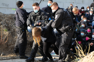 Eine Gruppe maskierter Personen in Mützen, einige bewaffnet, umgibt eine Frau, die von der Polizei eskortiert wird, mit Pflanzen, Steinen und einem protestbezogenen Wandtext im Hintergrund.