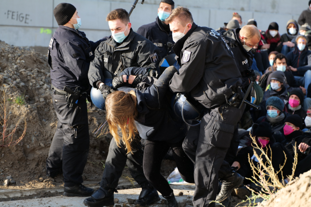 Eine Gruppe maskierter Personen in Mützen, einige bewaffnet, umgibt eine Frau, die von der Polizei eskortiert wird, mit Pflanzen, Steinen und einem protestbezogenen Wandtext im Hintergrund.