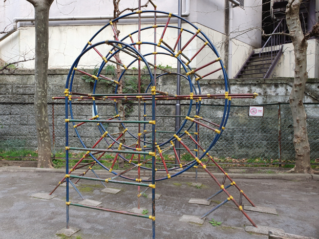 Spielplatz mit großem Metall-Klettergerüst, umgeben von Bäumen und einer Maschendrahtzaun, mit Treppen mit Geländern, die zu einem Gebäude mit Fenstern und Rohren führen, einem Schild an einem Pfahl und grasigem Boden.