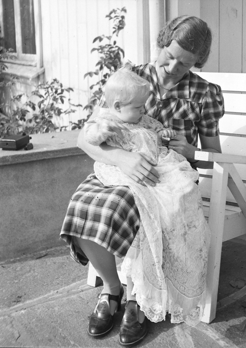 Eine Frau sitzt auf einer Bank und h├Ąlt ein Baby im Arm, beide teilen einen warmen, liebevollen Moment mit üppiger Vegetation und einem Fenster im Hintergrund.