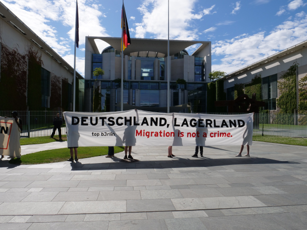 Eine Gruppe von Menschen, die ein Transparent mit der Aufschrift "Deutschland, Lagerland Migration ist kein Verbrechen" vor einem Fensterbau mit Fenstern, umgeben von Gras, einem Metallzaun, Pflanzen, Bäumen und Fahnen mit Stäben unter einem bewölktem Himmel halten.