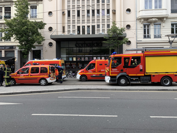 Eine Gruppe von Feuerwehrautos auf einer Pariser Straße geparkt, mit Menschen in der Nähe, vor Häusern, Bäumen und einem Fahrrad, wahrscheinlich nach einem Unfall.