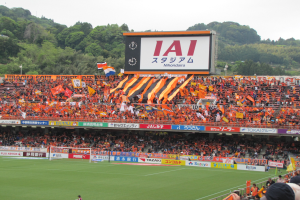 Ein Fußballspiel wird in einem Stadion mit einer großen Menge, grünem Rasen, einem Torpfosten, Bannern, Fahnen, einem großen Bildschirm, Bäumen und einem klaren blauen Himmel gespielt.