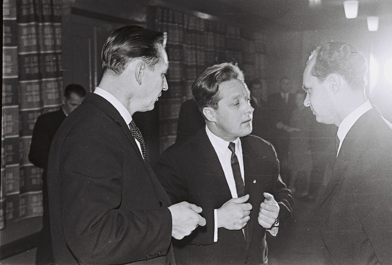 Drei Männer in Anzügen unterhalten sich in einem hell erleuchteten Raum mit Bücherregalen im Hintergrund, aufgenommen in Schwarz-Weiß.