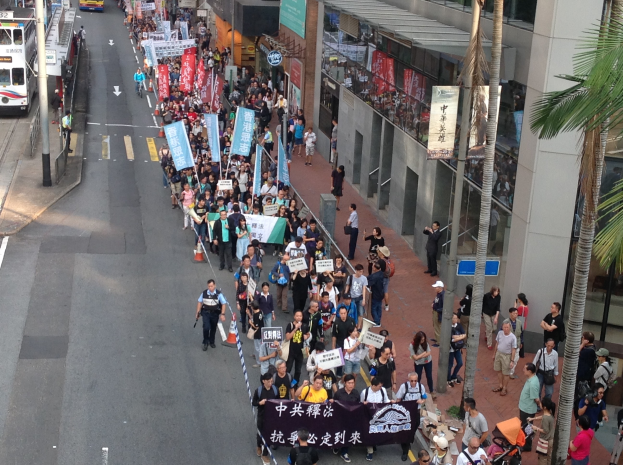 Eine große Gruppe von Menschen marschiert auf einer Straße in Hong Kong, trägt Schilder und Plakate, wobei Bäume, Glasgebäude, Fahrzeuge und Straßenschilder zu sehen sind.