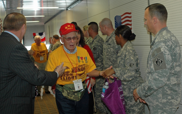 Eine Gruppe von Menschen und Soldaten, die drinnen mit einer Flagge an der rechten Wand und Lampen an der Decke stehen.