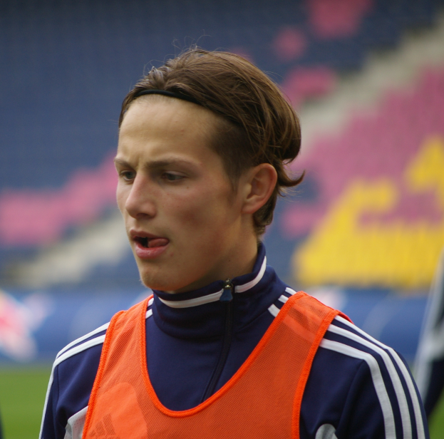 Ein junger Mann in einer orangen Weste steht auf einem Fußballfeld mit ausgestreckten Armen und entschlossener Miene, mit einem unscharfen Hintergrund.