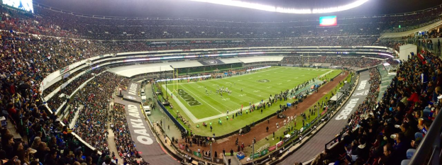 Ein großes Stadion voller Zuschauer, die ein Fußballspiel verfolgen, mit Spielern und verschiedenen Gegenständen auf dem Feld, beleuchteten Tafeln, Geländern, Bildschirmen und Deckenleuchten.