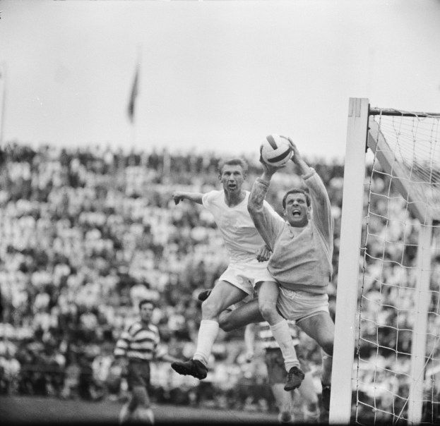 Zwei Männer beim Fußballspielen vor einem Publikum, mit einem Torpfosten auf der rechten Seite und einer Fahne im Hintergrund, in Schwarz-Weiß.