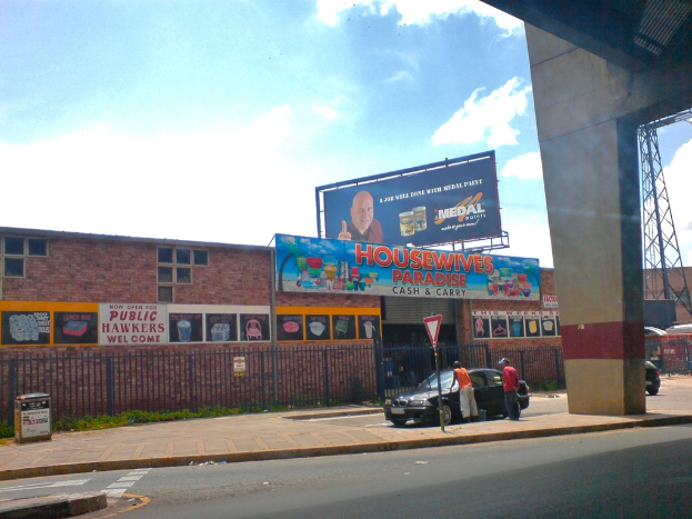 Eine Straße mit einer Säule und einem Auto auf der rechten Seite, ein Pfahl mit einer weißen und roten Tafel, zwei Männer neben einem Auto stehend, ein Müllcontainer auf der linken Seite, braune Gebäude mit Plakaten im Hintergrund, eine blaue Tafel mit Text oben auf einem Gebäude, eine blaue Werbetafel, ein Stromtransformator, ein rotes Objekt auf der rechten Seite und ein bewölkter Himmel darüber.