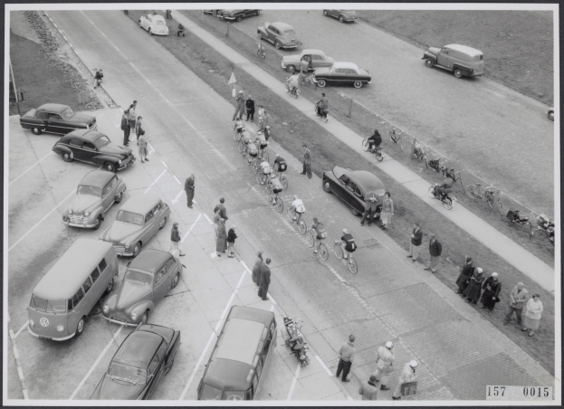 Schwarzes und weißes Foto einer belebten Straße mit Autos, Fußgängern und Fahrradfahrern, Gras am Straßenrand und Text unten.