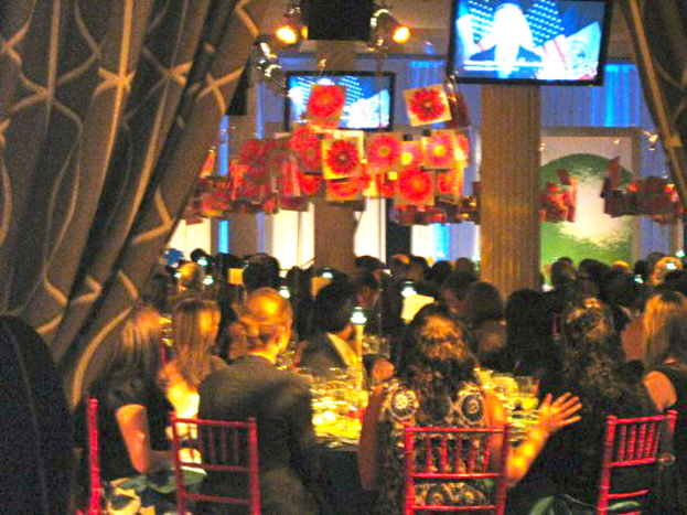 Eine Gruppe von Menschen sitzt an Tischen mit Essen, Fernsehern, Lampen, hängenden Papieren und Vorhängen in einem Raum.