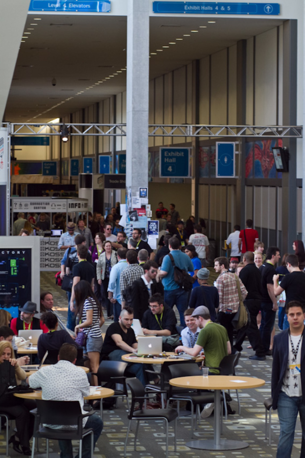 Ein belebter Airport-Lobby mit wartenden Menschen, einige stehen und andere arbeiten, Richtungsschilder an der rechten Wand und darüber sowie an einer Eisenstange an der Decke befestigte Lampen.
