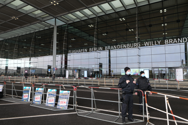 Zwei Polizeibeamte in schwarzen Uniformen stehen vor einem großen Glasgebäude mit Text darauf, mit Absperrungen und Schildern im Vordergrund und anderen Menschen und Gegenständen im Hintergrund.