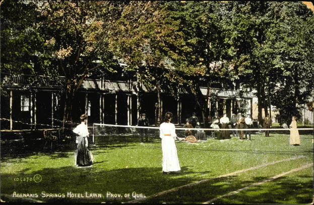 Altes Schwarz-Weiß-Foto einer Tennisveranstaltung auf dem Rasen des Abenakis Springs Hotel in Provo, Quebec, mit Spielern mit Schlägern, sitzenden Zuschauern, einem Netz, Bäumen und einem Gebäude im Hintergrund.