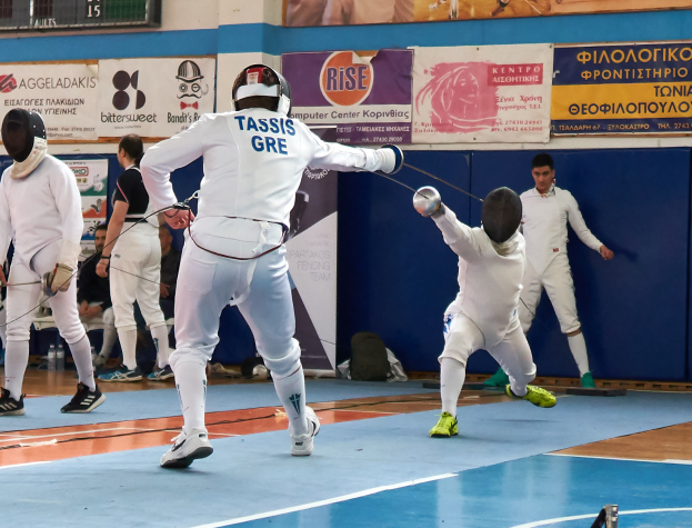 Eine Gruppe von Menschen in Helmen und mit Schwertern ficht in einer Turnhalle mit Bannern und einer Anzeigetafel im Hintergrund.