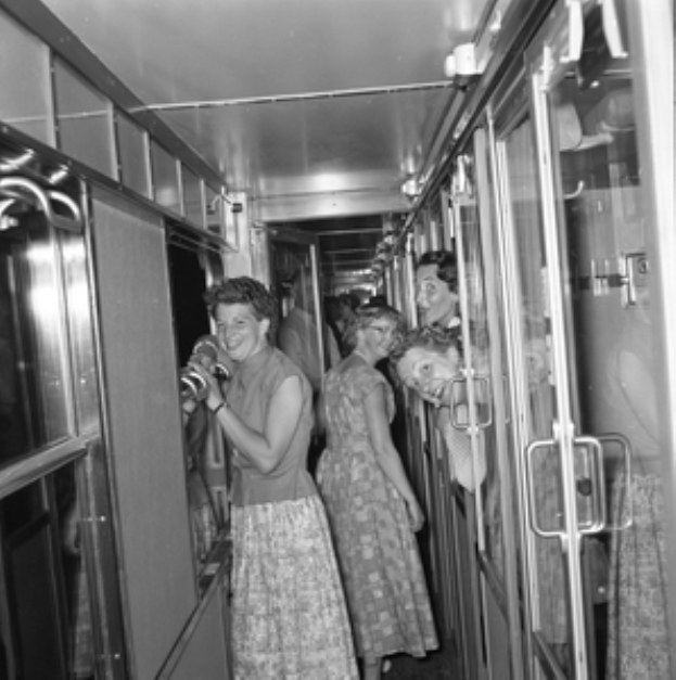 Eine Gruppe von Frauen, die auf einem Zug mit Türen auf beiden Seiten und Lampen oben steht, dargestellt in Schwarz-Weiß.