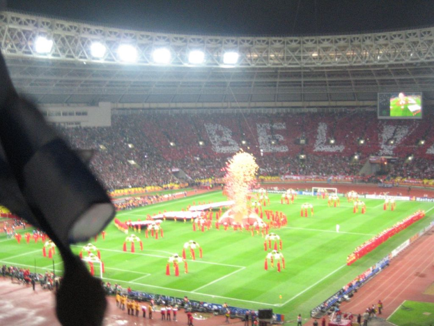 Ein Stadion mit Flutlicht, Eisengittern, Zuschauern, stehenden Personen, Werbetafeln, Anzeigetafeln, √úberwachungskameras und dekorativen Fahnen.