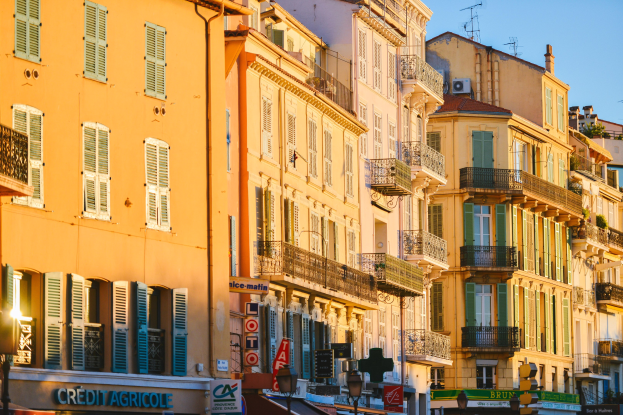 Eine Stadtstraße mit hohen Gebäuden, die Fenster, Balkone und Schilderzügen aufweist und von Bäumen gesäumt ist, unter einem klaren blauen Himmel.