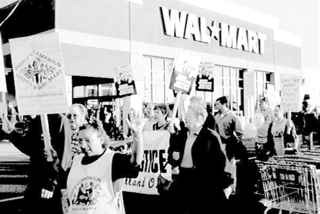 Ein Schwarz-Wei-Bild von Menschen, die protestieren und Plakate halten, mit einem Einkaufszentrum im Hintergrund.