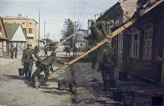 Gruppe von deutschen Soldaten im Zweiten Weltkrieg in militärischer Uniform auf einer Straße neben einem Gebäude stehend, mit Helmen und hölzernen Gegenständen mit Eimern auf dem Boden, umgeben von Gebäuden, Bäumen, Pfählen und Drähten unter einem klaren Himmel.