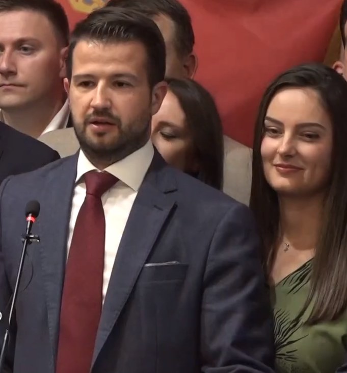 Ein Mann im Anzug spricht an einem Rednerpult mit einem Mikrofon zu einer Menge, während im Hintergrund ein Banner hängt.