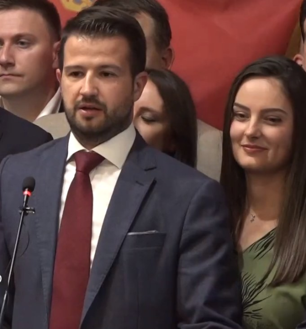 Ein Mann im Anzug spricht an einem Rednerpult mit einem Mikrofon zu einer Menge, während im Hintergrund ein Banner hängt.